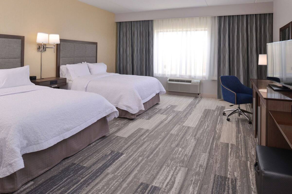 Une chambre d’hôtel avec deux lits, un bureau avec une chaise, une télévision et une fenêtre avec des rideaux.