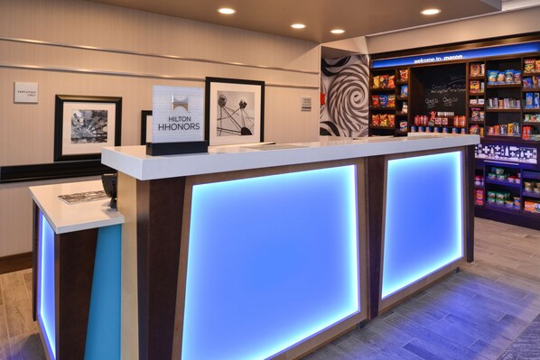 A reception area with a modern design, featuring a reception desk, framed artwork, and a snack bar with various products. - Hampton Inn & Suites Cincinnati-Mason (Mason)