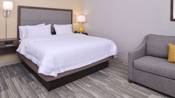 Una cama bien hecha con sábanas blancas, un cabecero tapizado de gris y un sofá color gris con un cojín estampado.