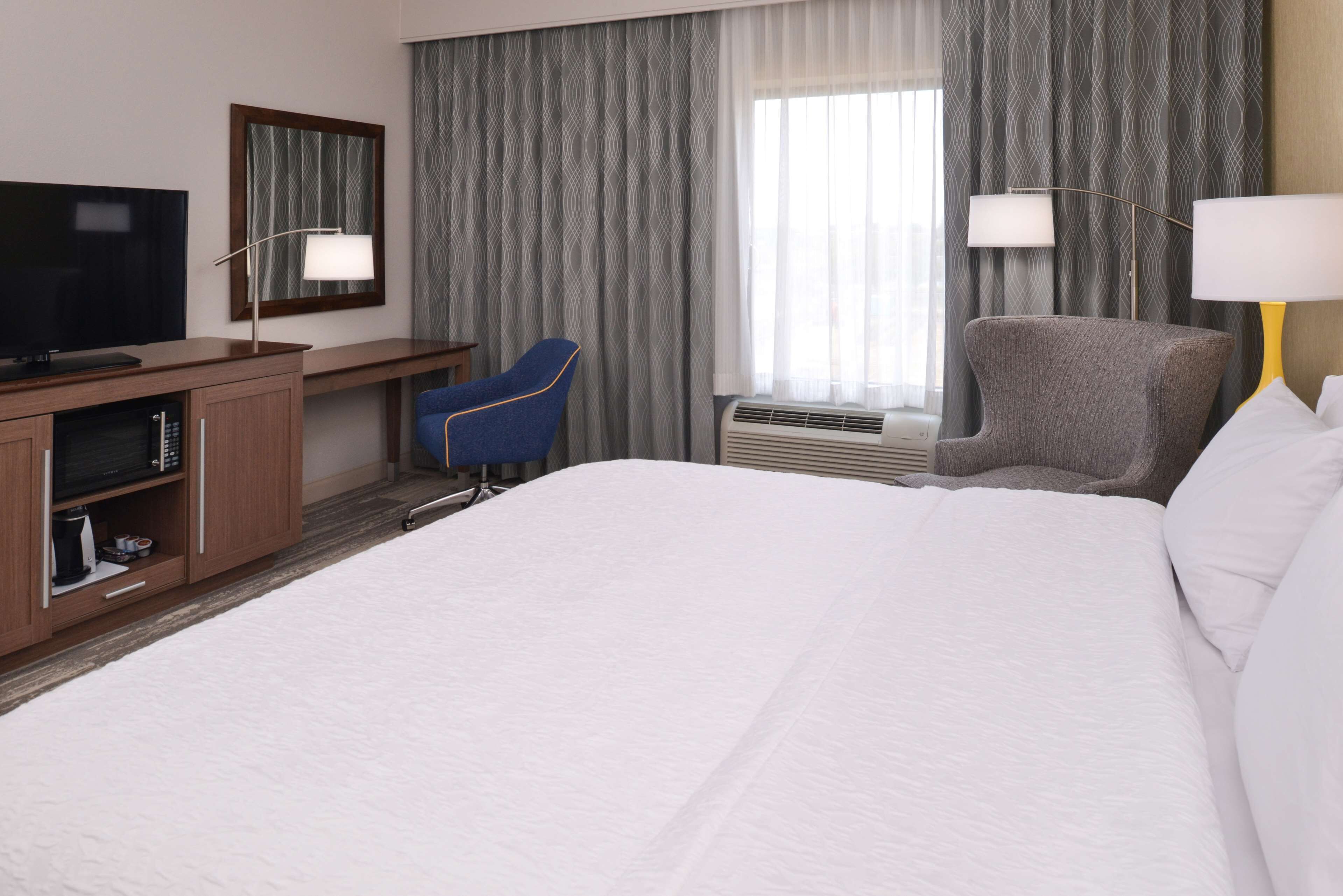 A hotel room with a large bed, a desk with a flat-screen TV, a blue chair, a grey armchair, and two bedside lamps.