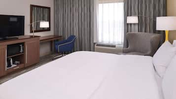 A hotel room with a large bed, a desk with a flat-screen TV, a blue chair, a grey armchair, and two bedside lamps.
