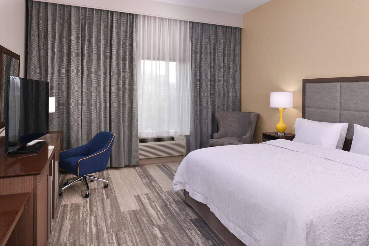 Une chambre d’hôtel avec un lit, un bureau avec une télévision, une chaise, une lampe et une fenêtre avec des rideaux.