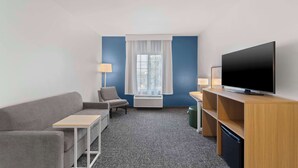 A hotel room with a grey sofa, a small table, a TV on a wooden stand, a lamp, and a window with curtains. - Spark by Hilton San Antonio Dominion (San Antonio)