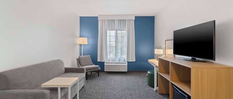 Habitación de hotel con sofá gris, una mesita, un televisor sobre un mueble de madera, una lámpara y una ventana con cortinas.