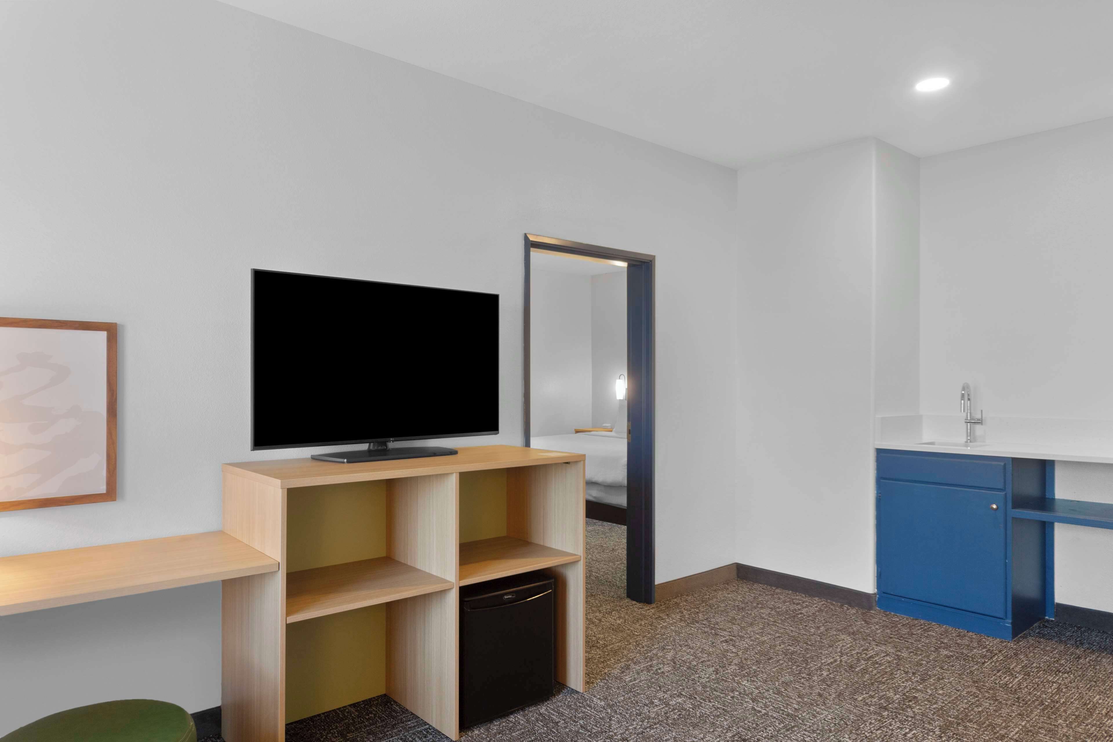 Habitación de hotel moderna con televisión de pantalla plana, un escritorio de madera con estantes, un refrigerador pequeño, un armario azul y un espejo.
