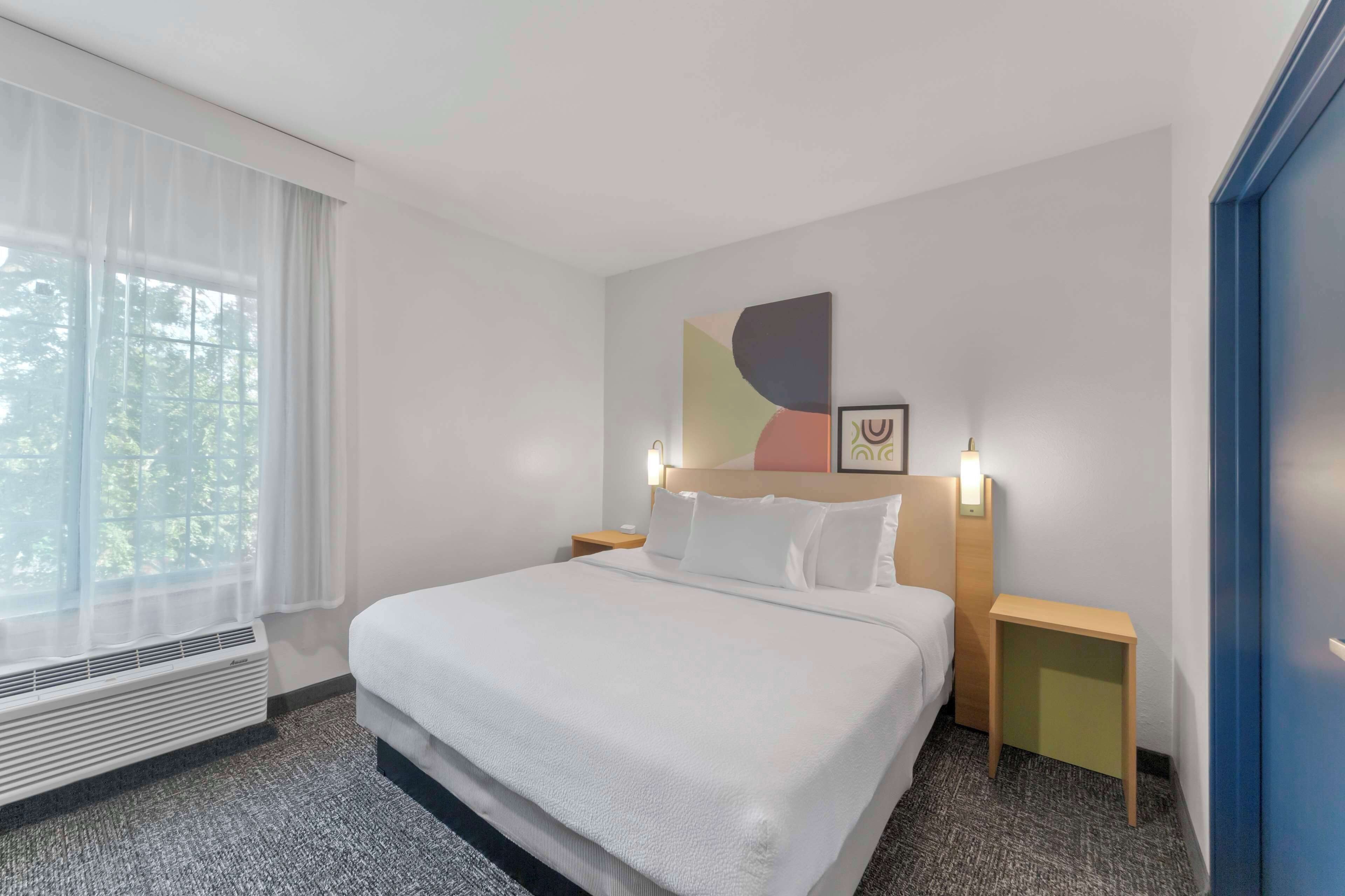 Ein Hotelzimmer mit einem großen Bett, einem Nachttisch, einem Wandbild und einem Fenster mit transparenten Vorhängen.