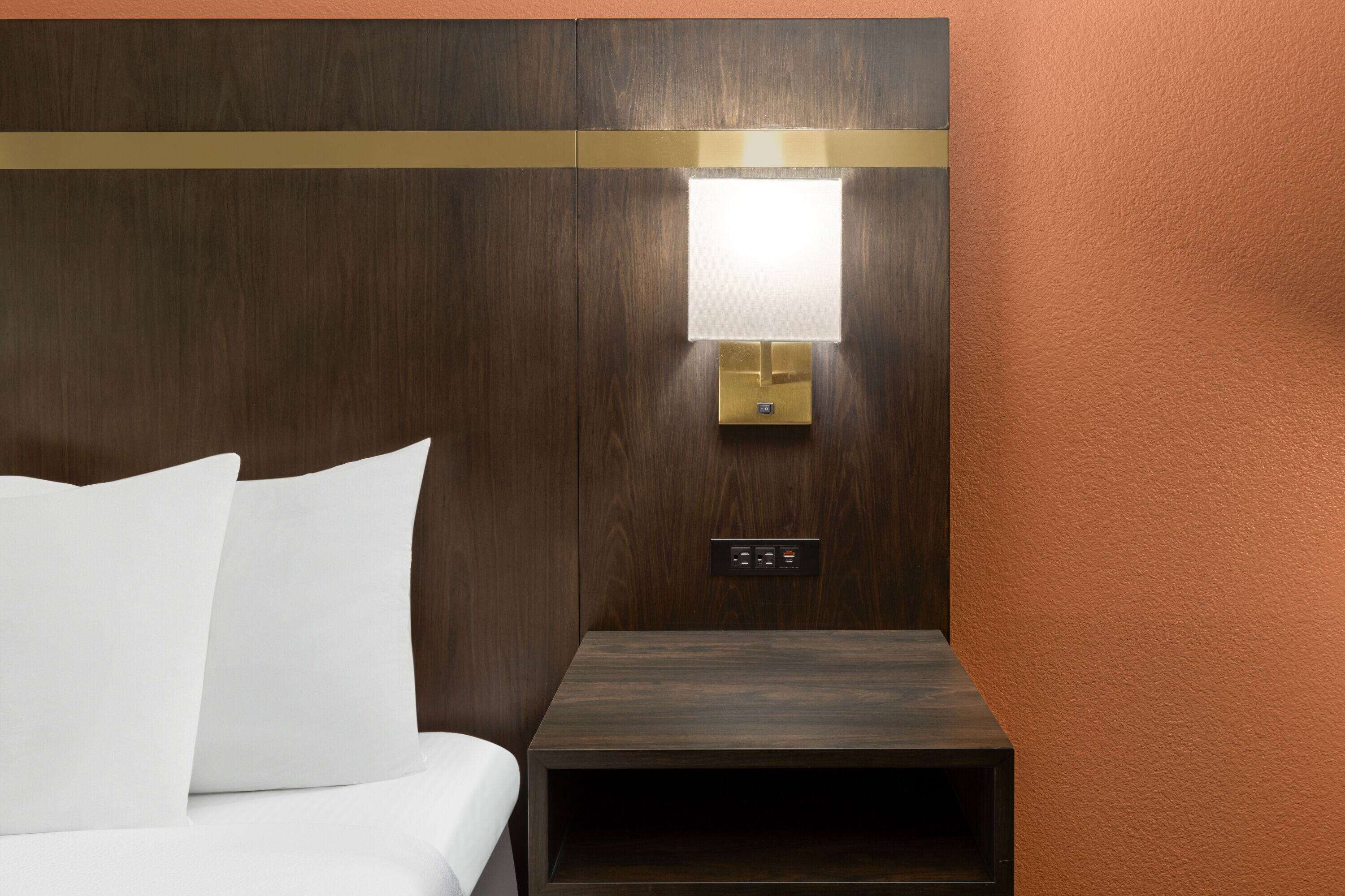A hotel room with a bed, a nightstand, a wall-mounted lamp, and a control panel.