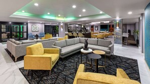 A modern hotel lobby with a sectional sofa, yellow armchairs, a round table, and a bar area in the background. - La Quinta Inn & Suites by Wyndham Olathe (Olathe)