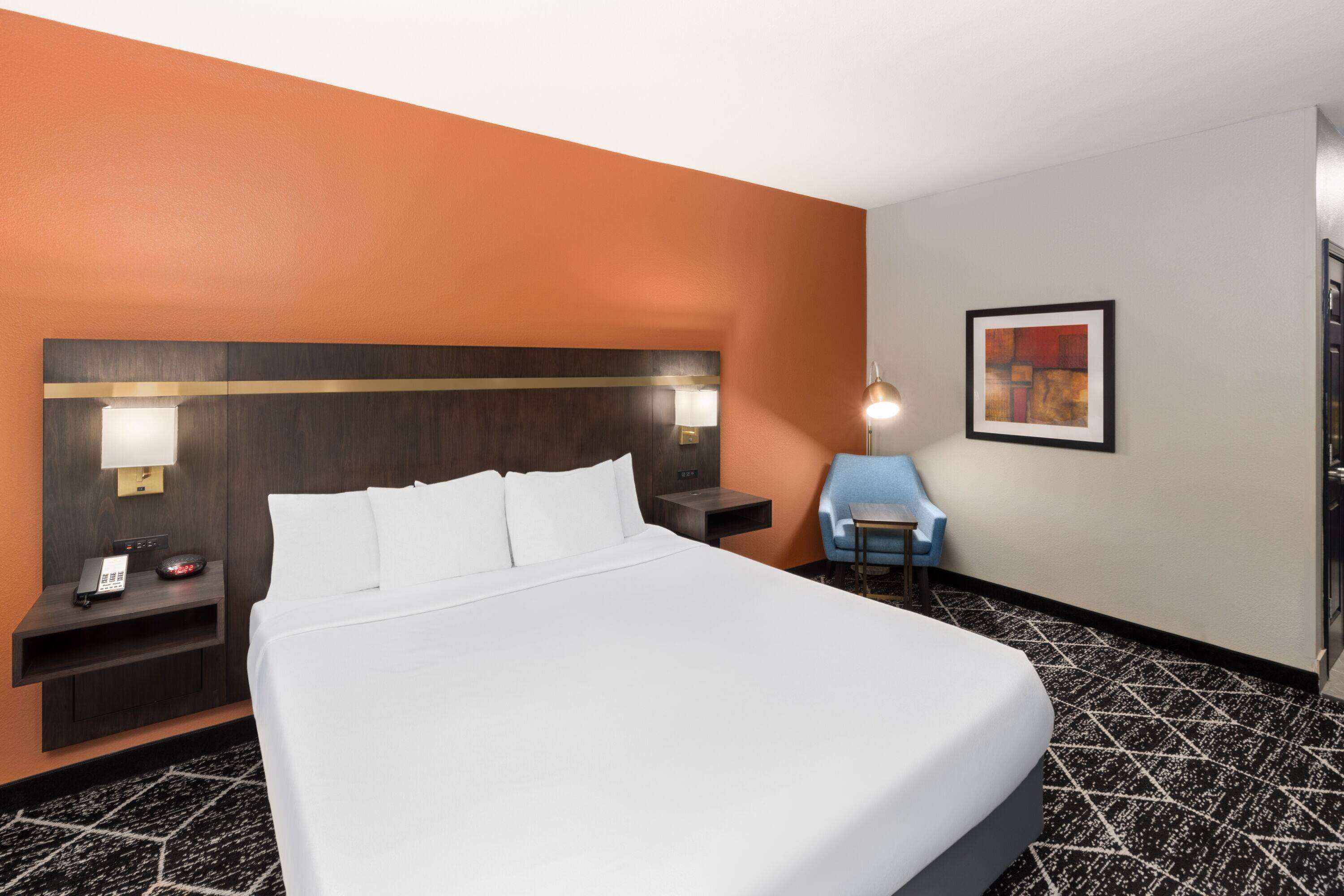 A hotel room with a large bed, a nightstand with a phone and lamp, a small table with a chair, and a framed picture on the wall.