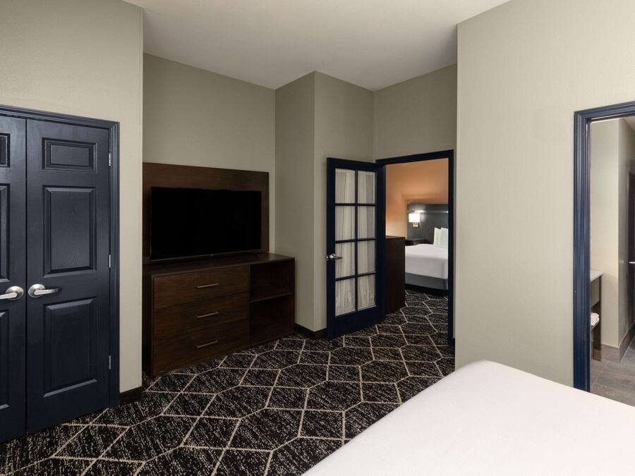 Una habitació d'hotel amb un llit gran, una televisió, un taulell i una porta que dona a una altra habitació.