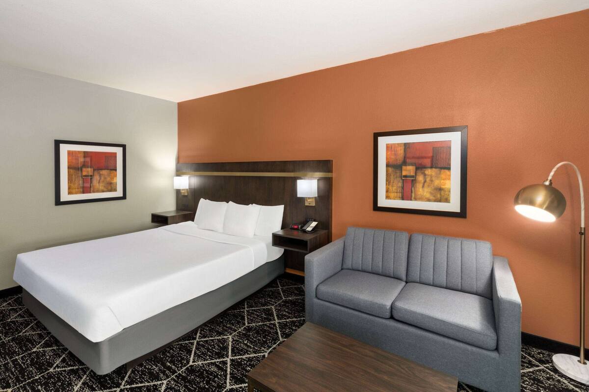 A hotel room with a bed, a gray sofa, a nightstand with a lamp, and two framed pictures on the walls.