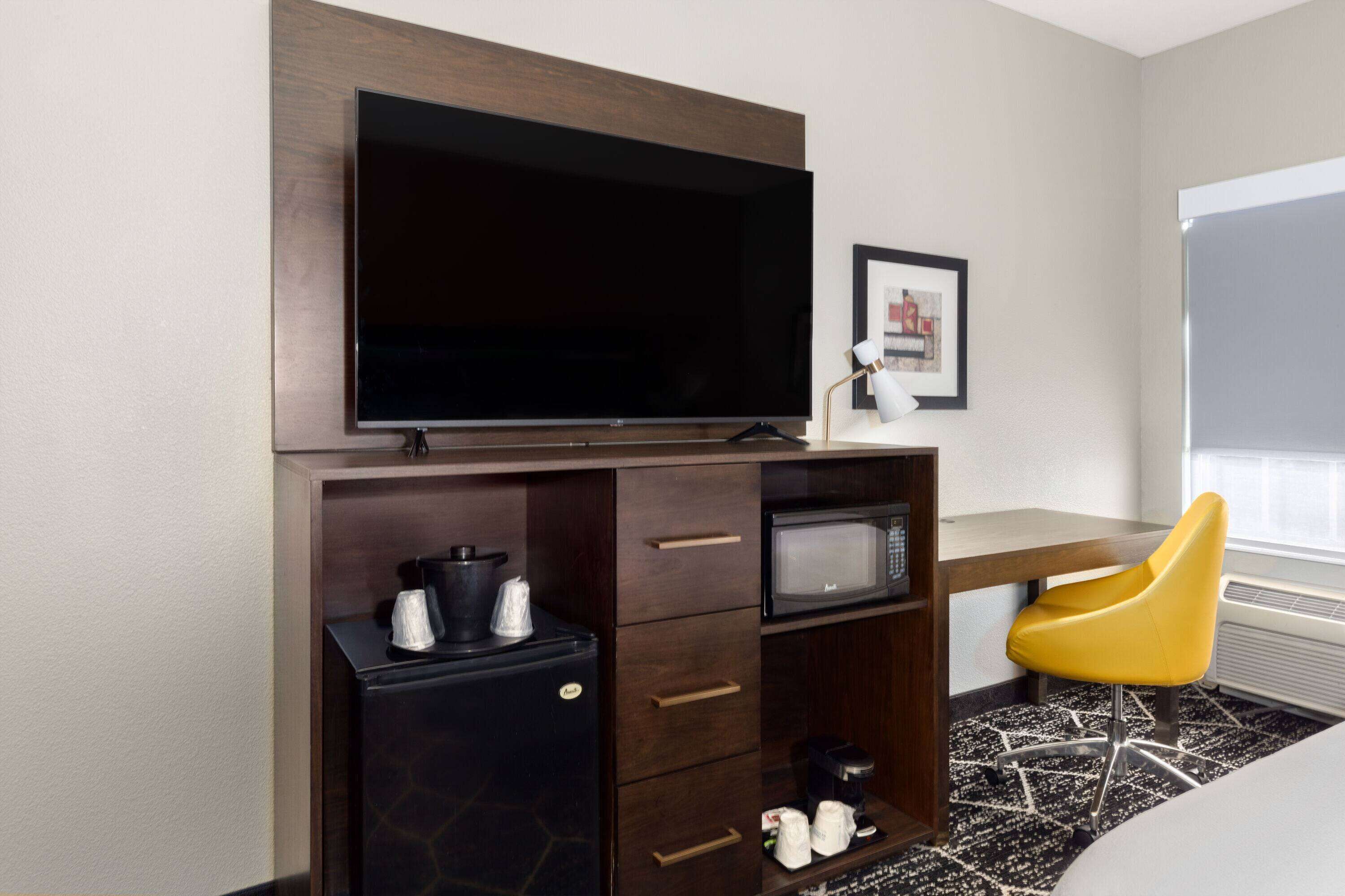 Habitación de hotel con televisor de pantalla plana montado en un mueble de madera, microondas, refrigerador pequeño, una silla amarilla y un cuadro enmarcado en la pared.