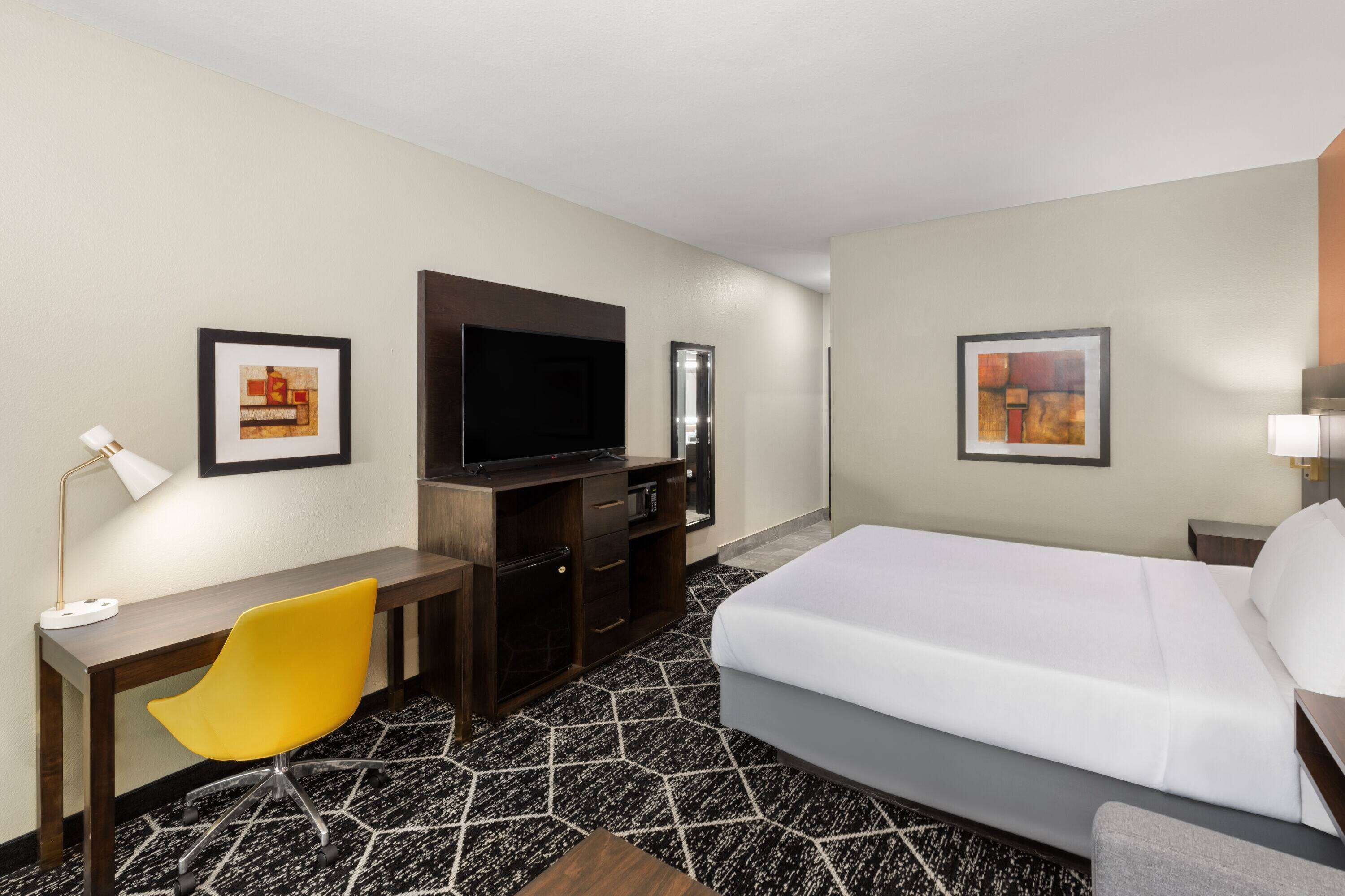 A hotel room with a bed, a desk, a chair, a TV, and two framed pictures on the walls.