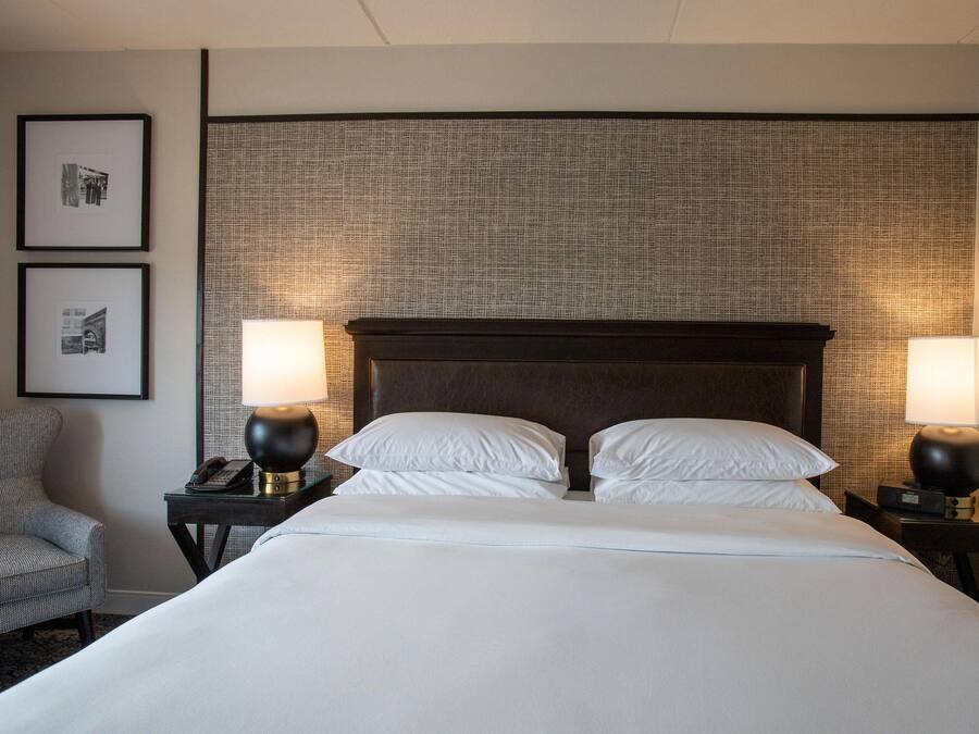 Habitación de hotel con una cama grande, dos mesitas de noche con lámparas, una silla y cuadros en la pared.