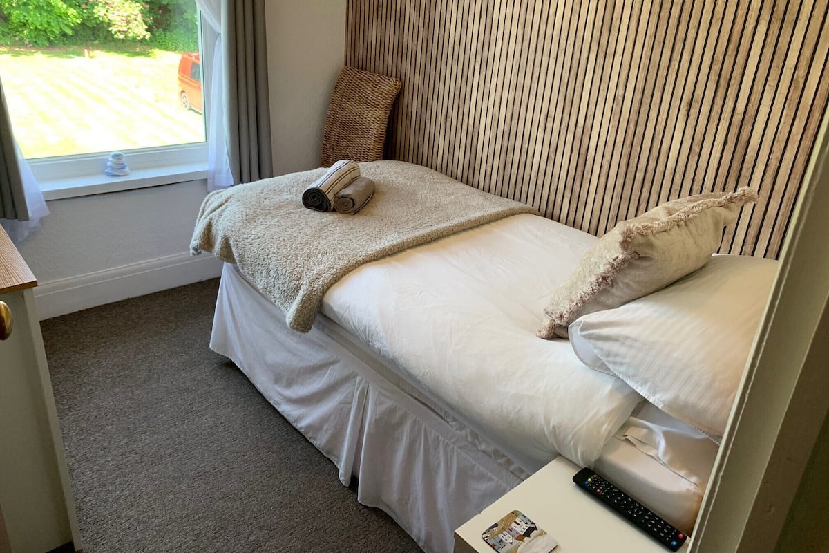 A neatly made bed with a rolled towel, a remote control, and a book on a bedside table.