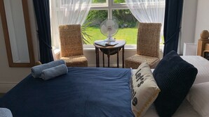 A bedroom with a bed, two wicker chairs, a small table with a fan, and a view of a garden through sheer curtains.