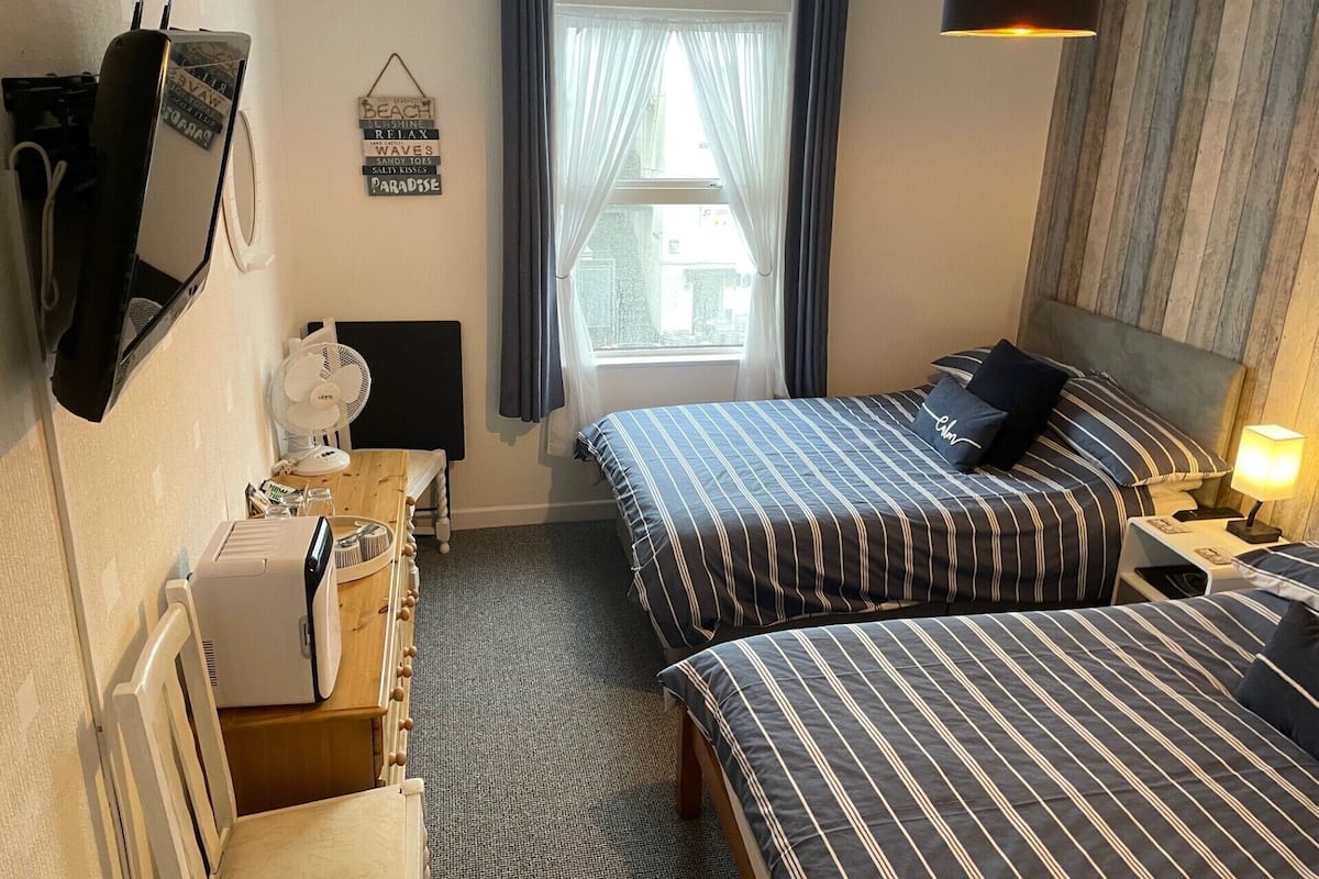 A hotel room with two beds, a desk with a computer, a fan, and a window with curtains.