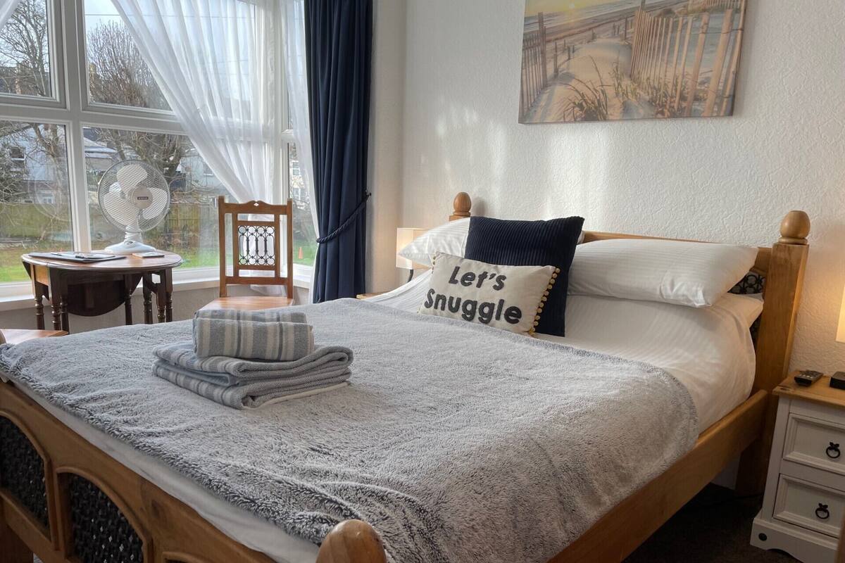 A neatly made bed with a "Let's Snuggle" pillow, a bedside table with a lamp, and a chair by a window with sheer curtains.