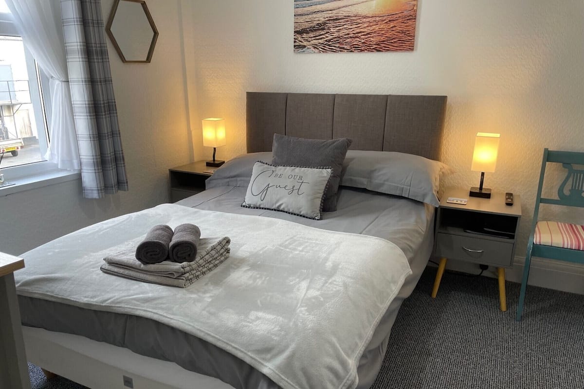 A neatly made bed with a pillow, a bedside table with a lamp, and a chair.
