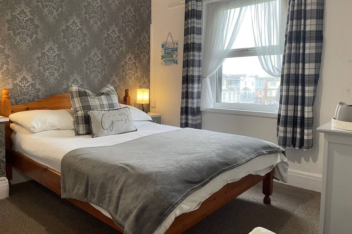 A neatly made bed with a grey blanket and a pillow with the text "BE OUR GUEST".