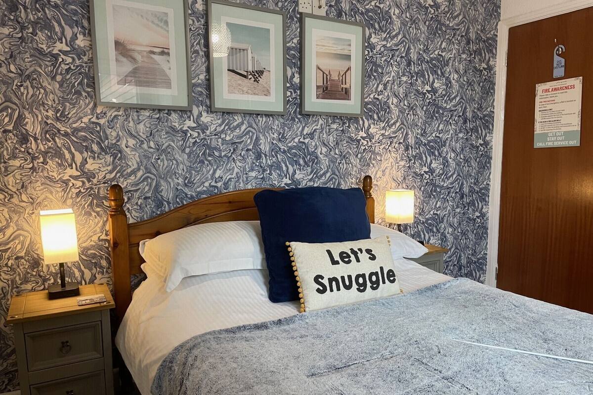 A neatly made bed with a pillow that reads "Let's Snuggle" and a blue throw blanket. There are three framed pictures on the wall above the bed, and a fire awareness sign on the door.