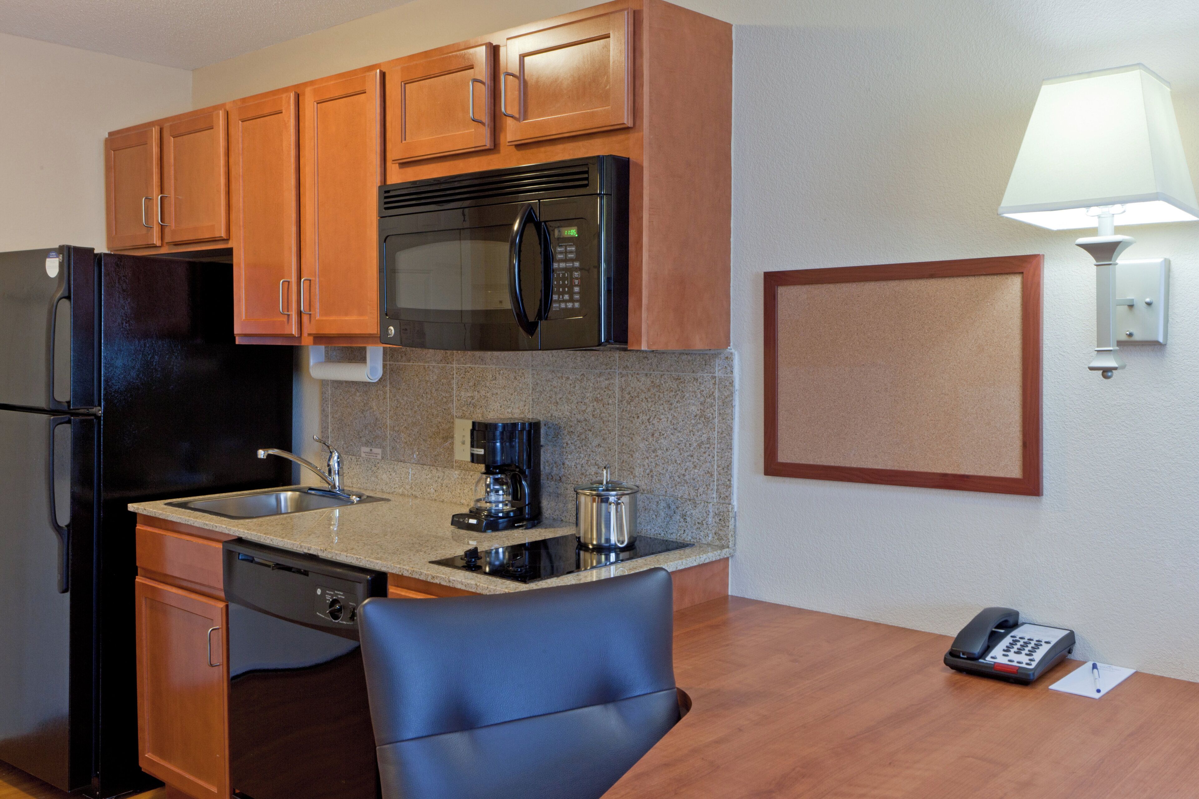 A compact hotel kitchen with wooden cabinets, a stainless steel refrigerator, a microwave, a coffee maker, and a telephone.