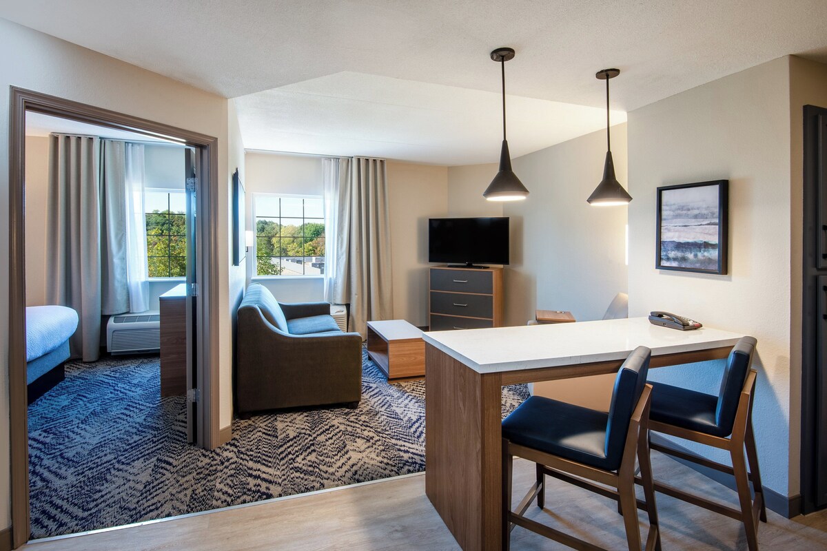 A modern hotel room with a sofa, a small dining area, a TV, and a view of greenery outside.