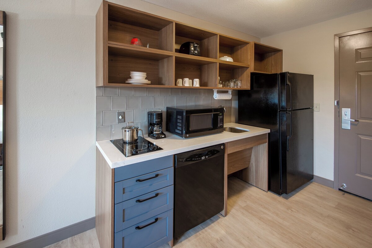 A compact kitchen with a dishwasher, microwave, refrigerator, and wooden shelves above the counter.