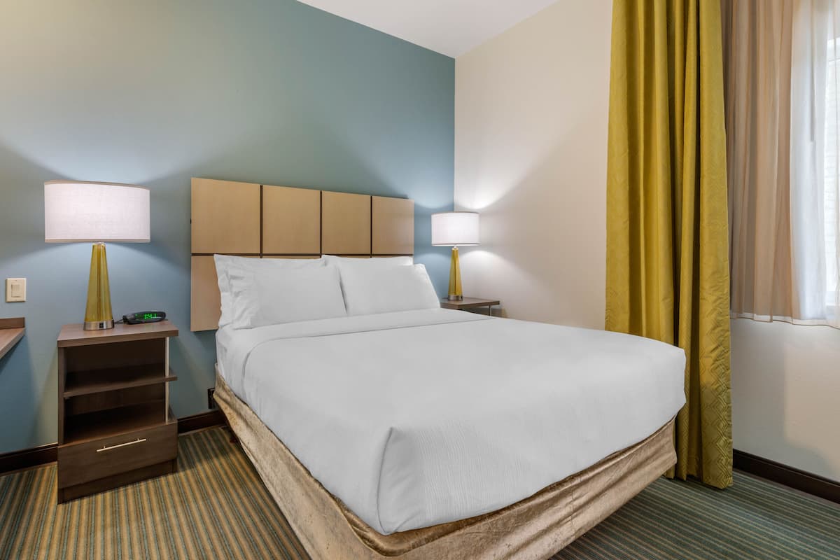 Une chambre d’hôtel avec un grand lit, deux tables de chevet avec des lampes, une petite table et une fenêtre avec des rideaux.
