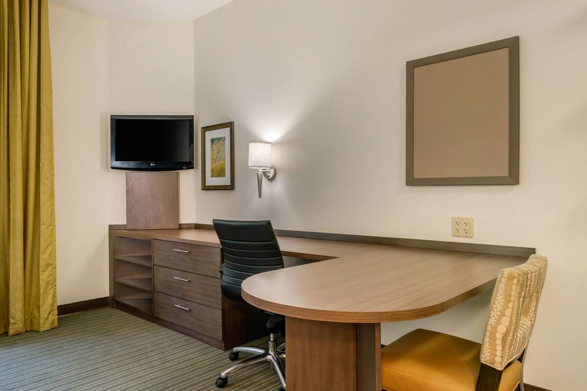 Une chambre d’hôtel avec un bureau en bois, un fauteuil de bureau noir, un téléviseur fixé au mur, un tableau encadré et une lampe fixée au mur.