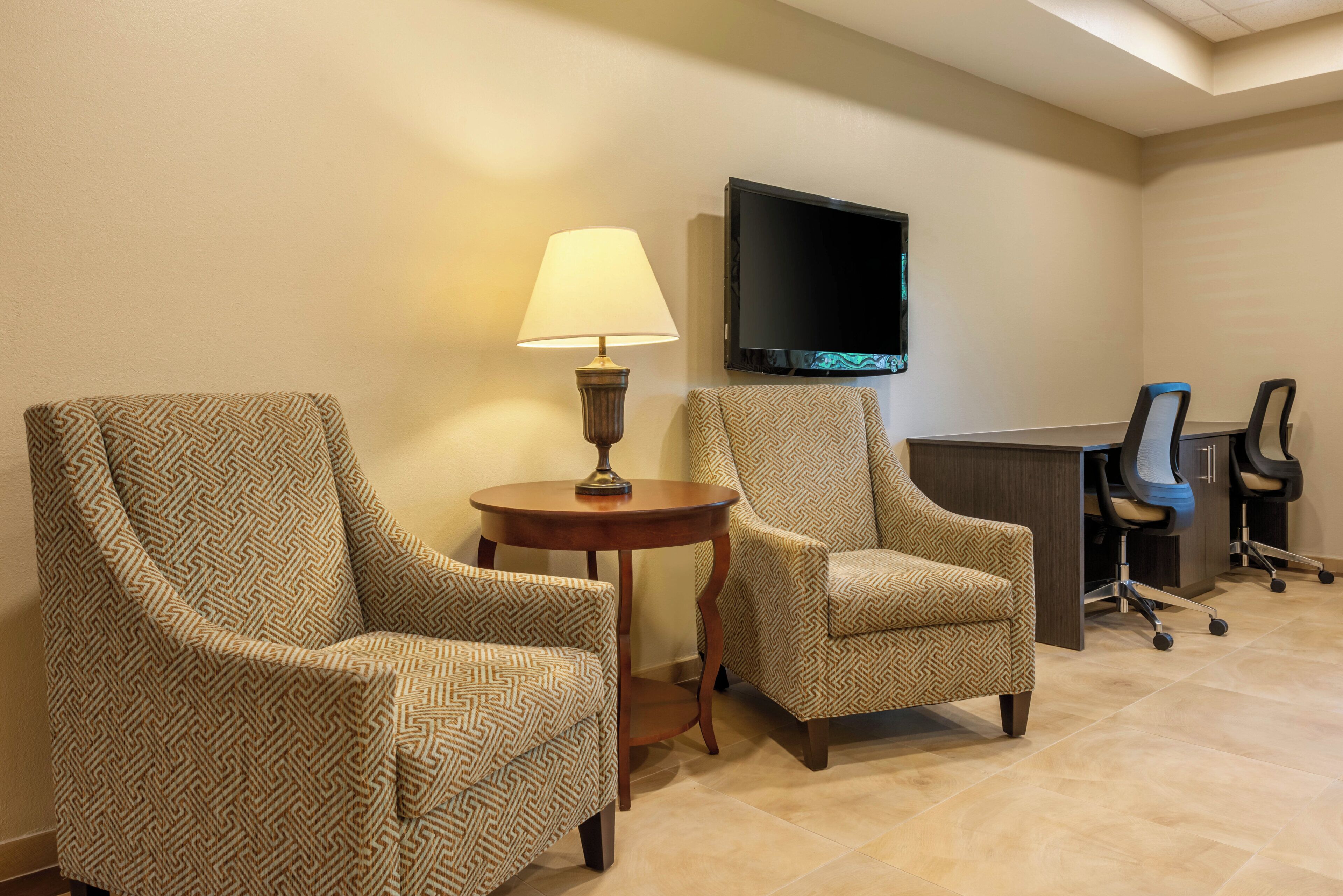 A hotel room with two armchairs, a small round table with a lamp, a flat-screen TV mounted on the wall, and a desk with office chairs.
