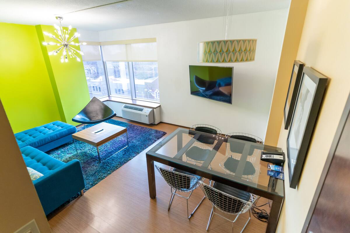 A modern living room with a glass dining table, blue sofa, and a large window with a view.