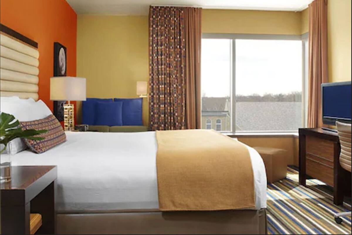 A hotel room with a large bed, a desk, a chair, a TV, and a window with a view of buildings.