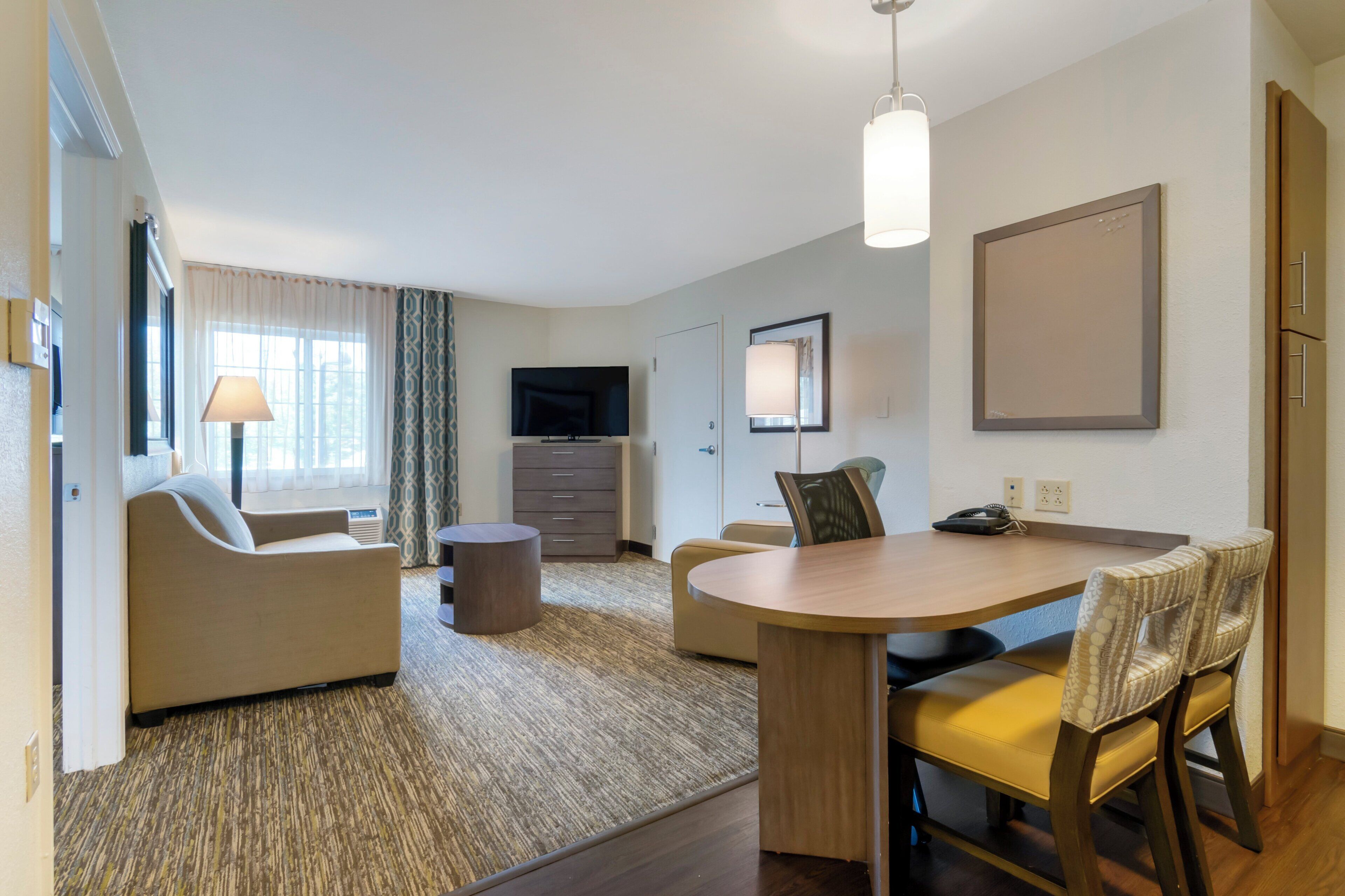 Ett modernt hotellrum med en soffa, ett matbord med stolar, en TV och ett stort fönster med gardiner.