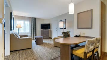 Ett modernt hotellrum med en soffa, ett matbord med stolar, en TV och ett stort fönster med gardiner.