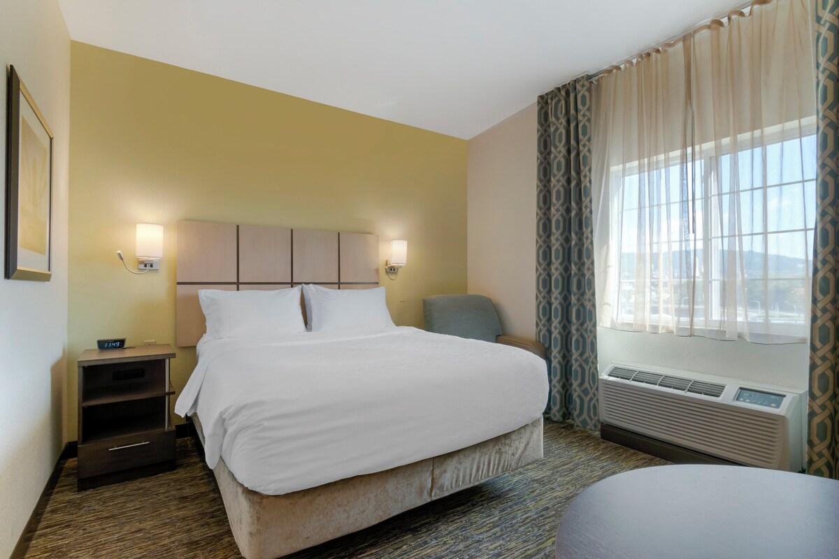 A hotel room with a bed, bedside tables, a chair, a window with curtains, and a wall-mounted air conditioner.