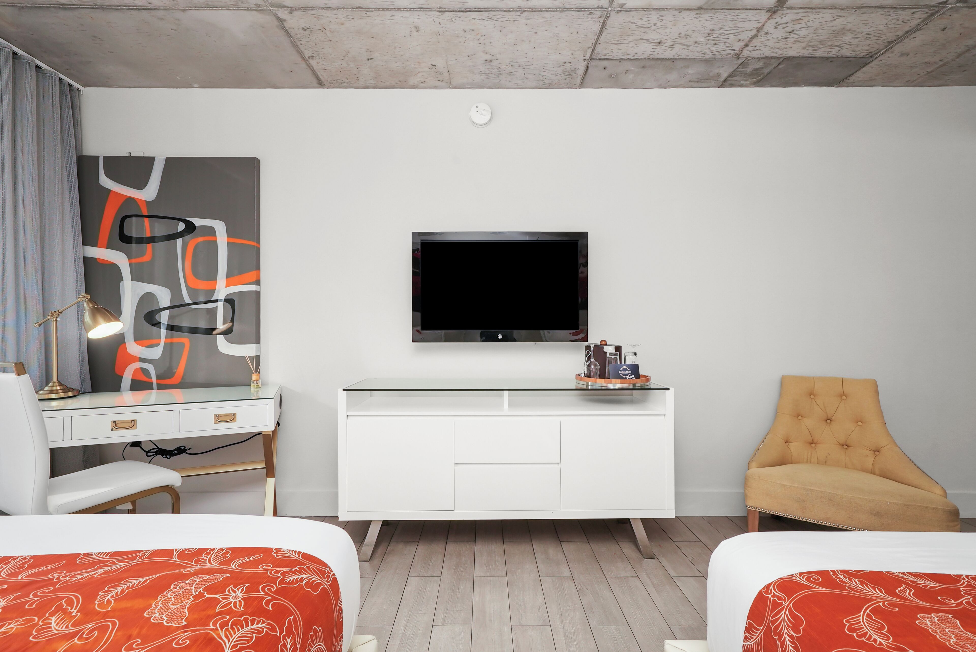 Ein modernes Hotelzimmer mit weißem Schreibtisch, einem Flachbildfernseher, einem bequemen Sessel und zwei Betten mit floralem, rotem Überwurf.