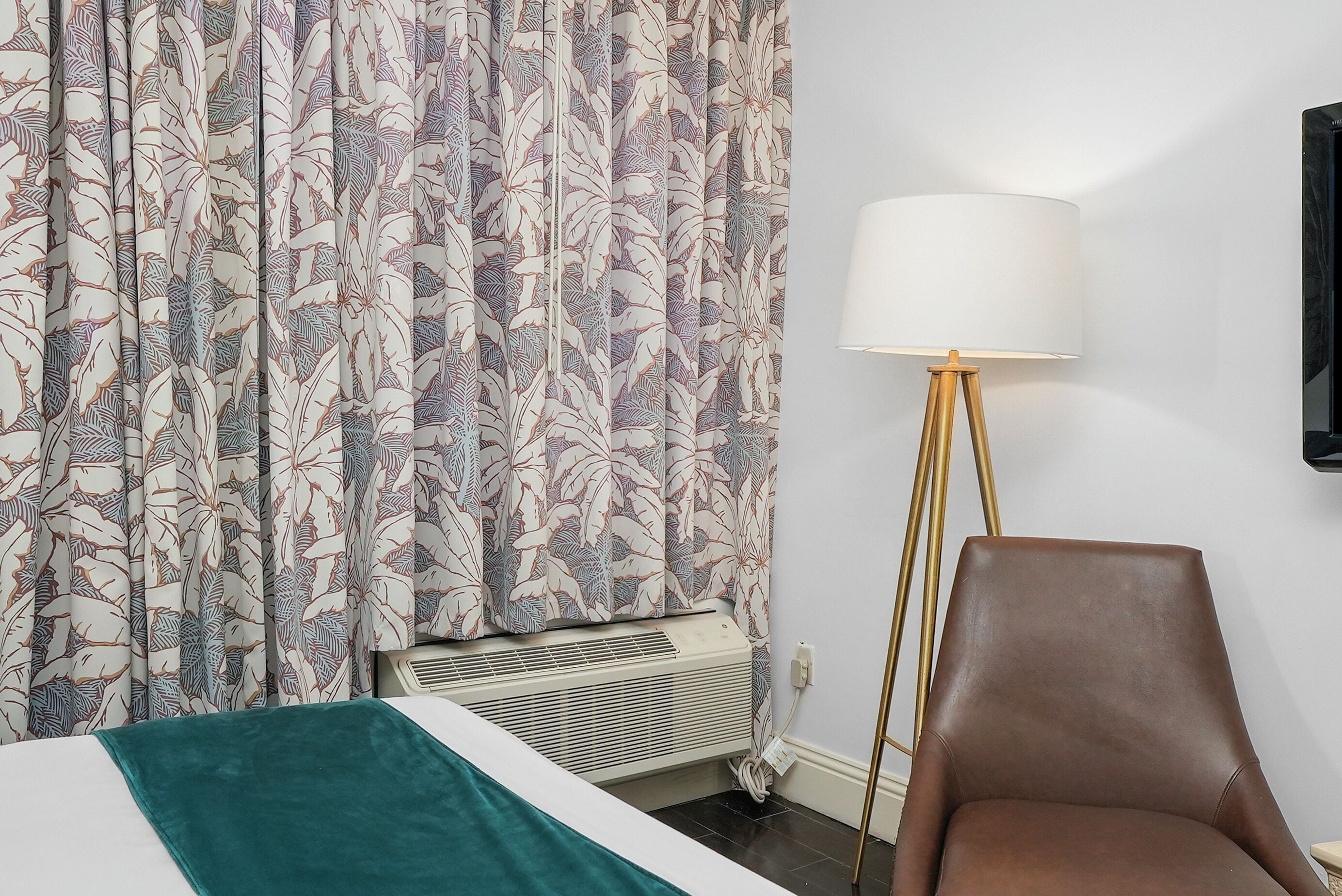 Una habitación de hotel con una cama, una manta verde, un sillón de cuero marrón, una lámpara de pie y un televisor montado en la pared.