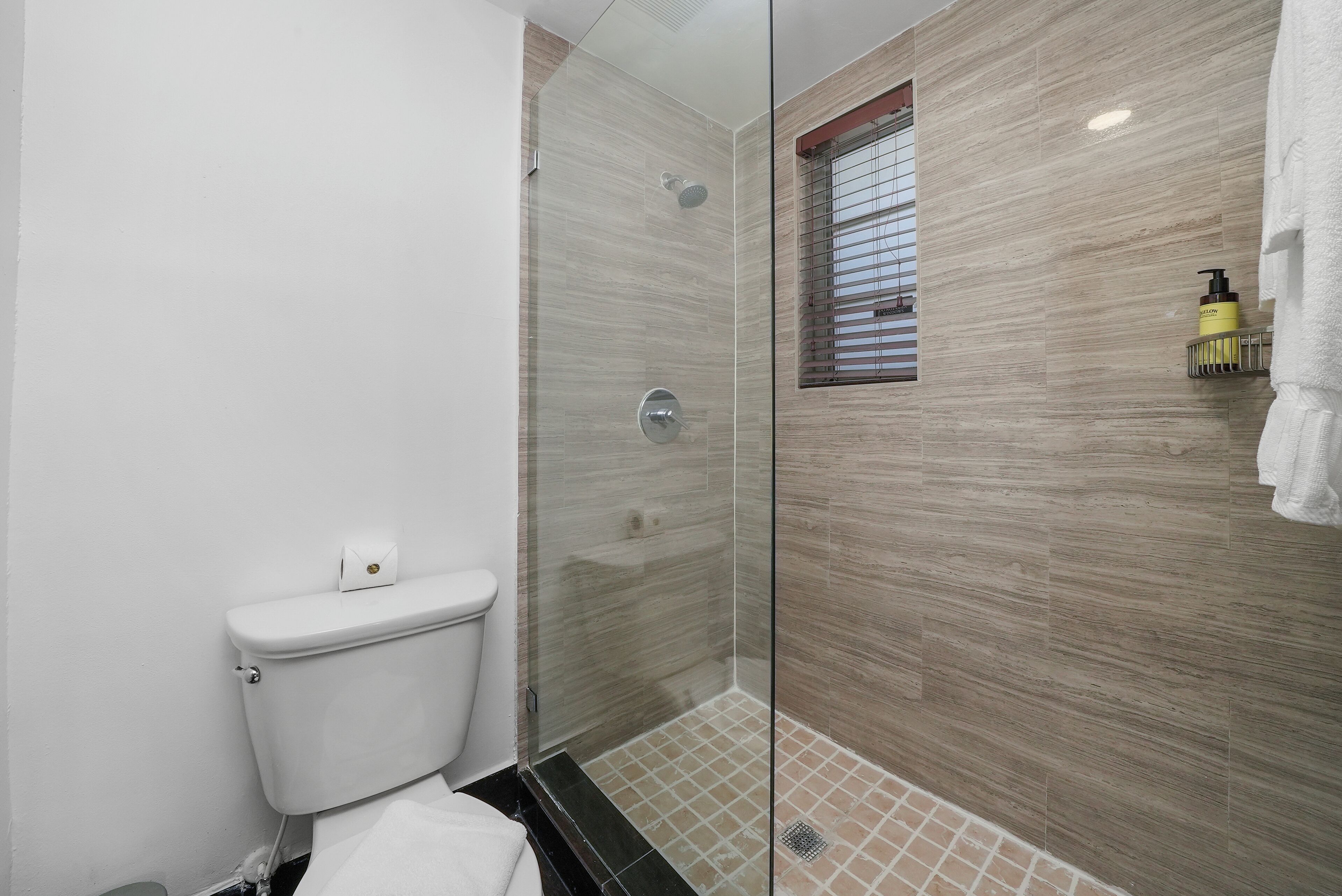 A modern bathroom with a glass-enclosed shower, a toilet, and a small window with blinds.