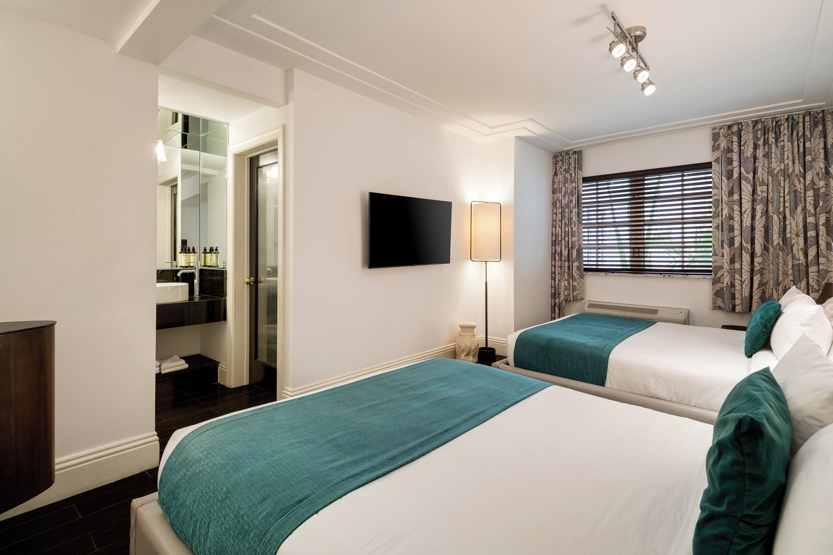 A hotel room with two beds, a TV, a lamp, a window with curtains, and a bathroom visible through an open door.