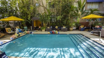 A swimming pool with lounge chairs and umbrellas, surrounded by greenery and a building.