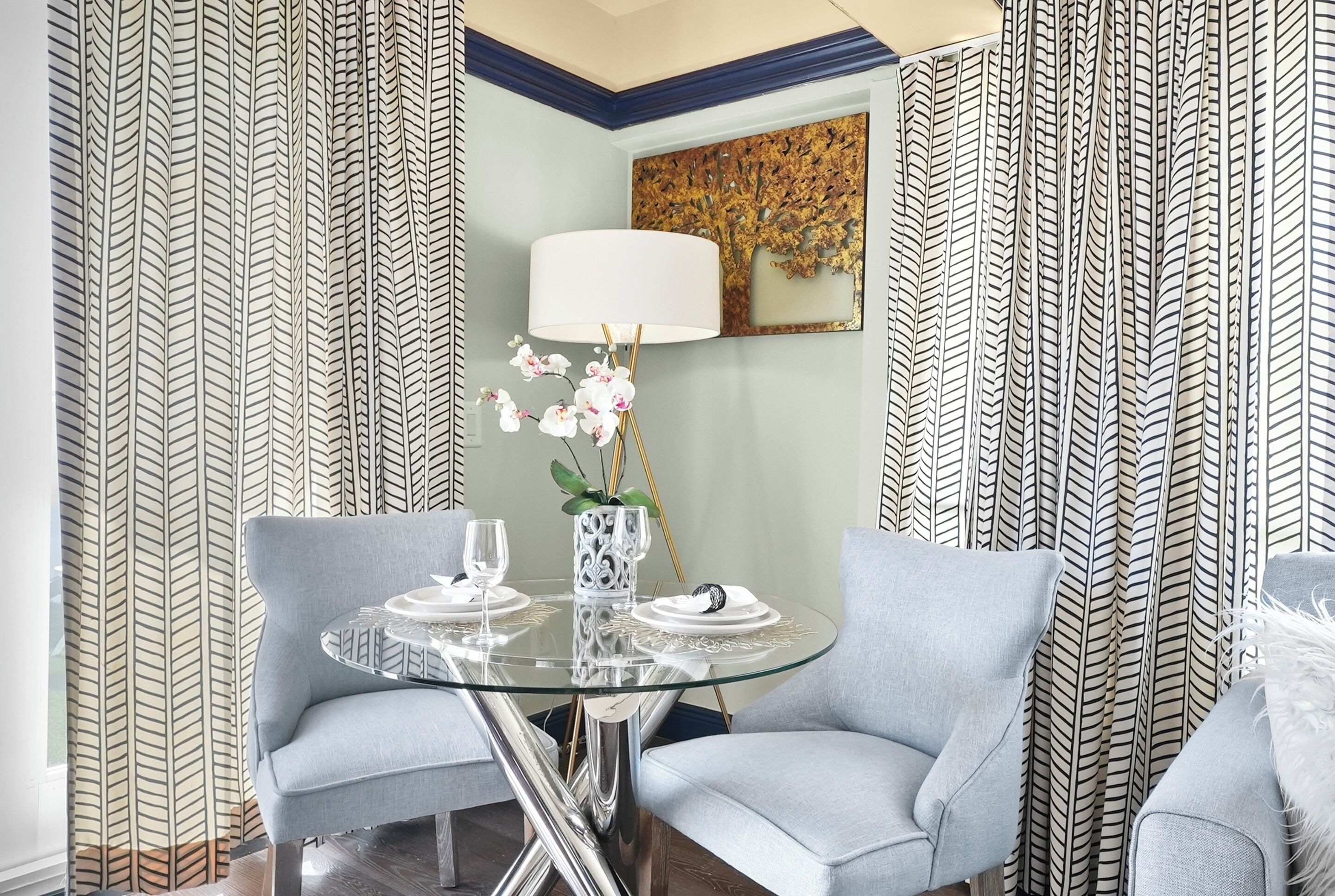 A modern dining area with a glass table, two grey upholstered chairs, a white table lamp, and a decorative vase with orchids.