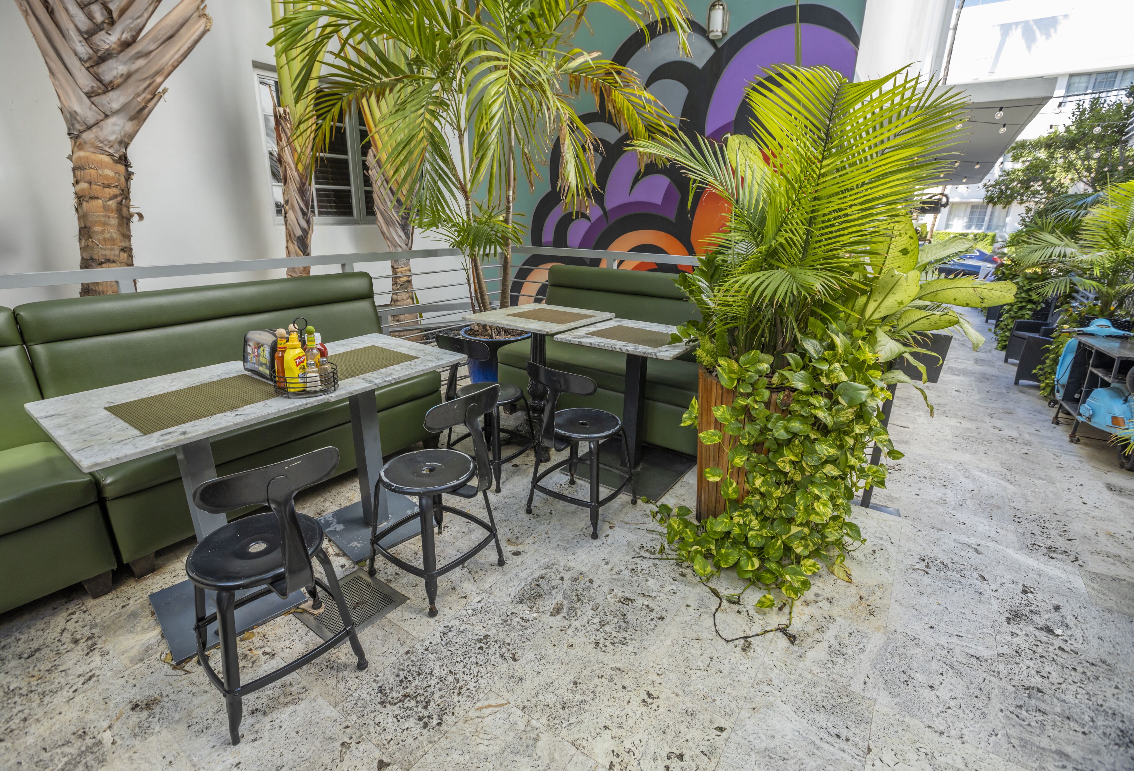 A green outdoor seating area with a table and chairs, surrounded by potted plants and a mural.