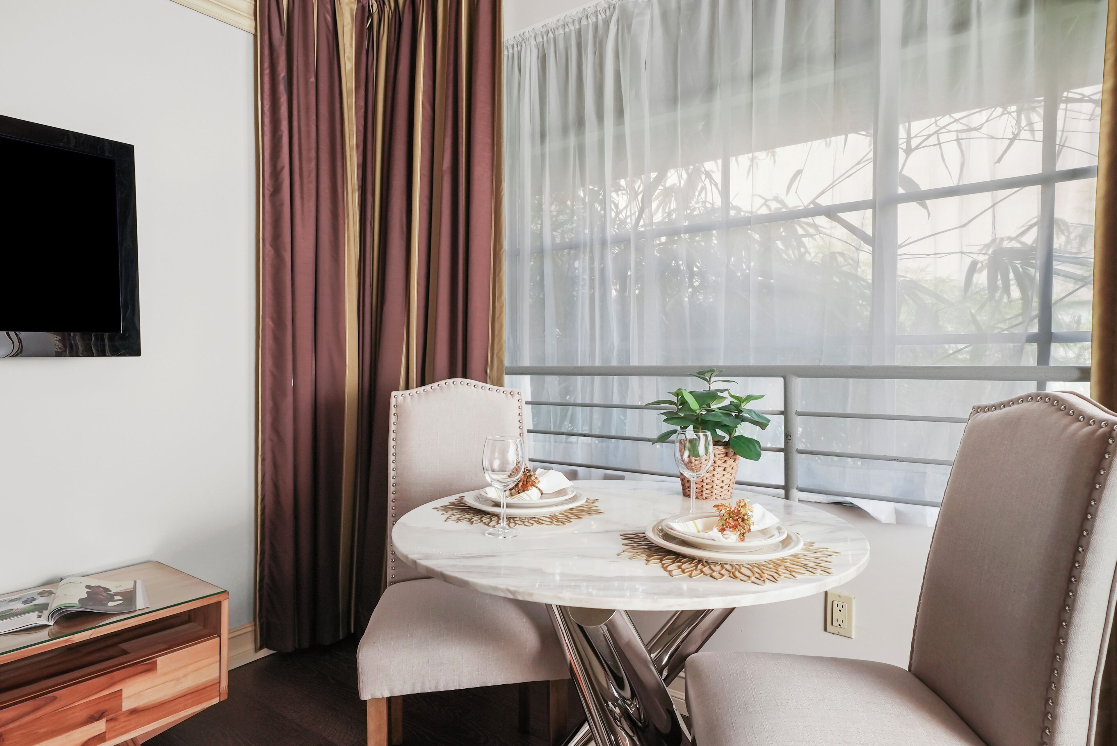 A dining area with a round table, two chairs, a glass-top side table, a television on the wall, and a window with curtains.