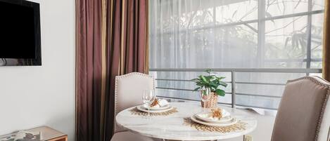 A dining area with a round table, two chairs, a glass-top side table, a television on the wall, and a window with curtains.