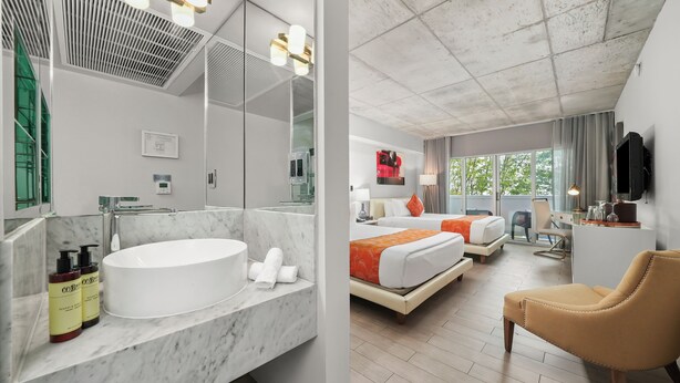 A modern bathroom with a large sink and mirror, a shower area, and toiletries. The room has a bed with orange bedding, a desk, and a chair. - Riviera Hotel South Beach (Miami Beach)
