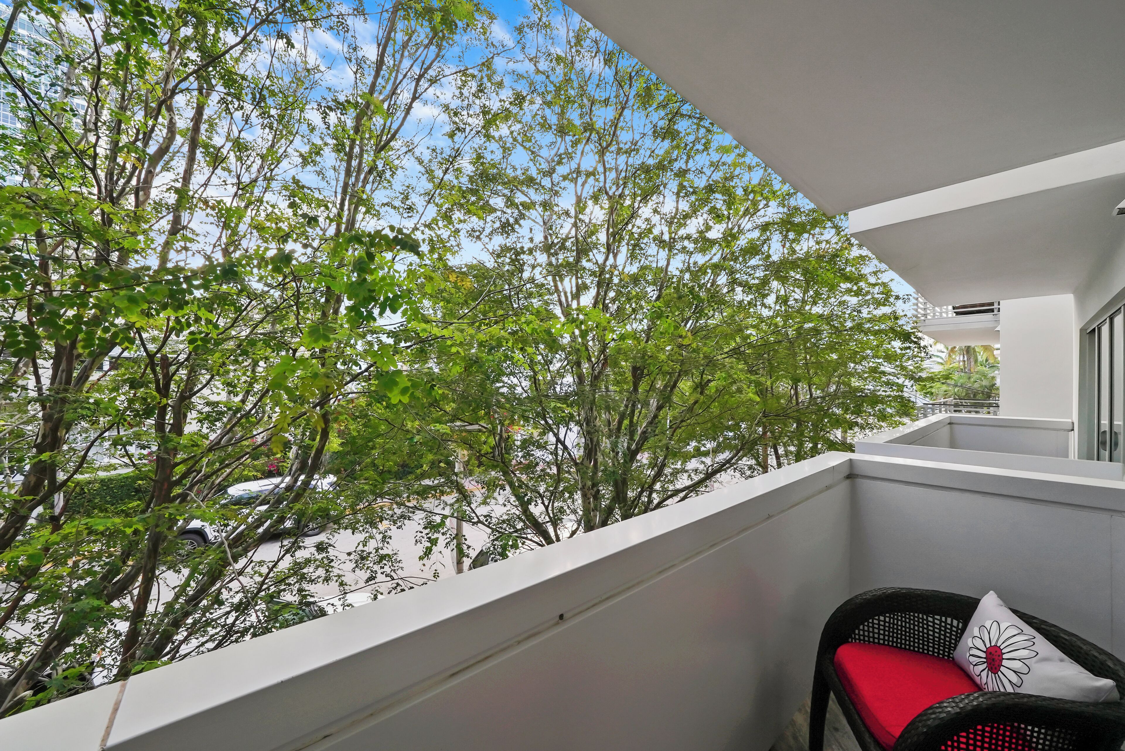 A balcony with a wicker chair and a red cushion, overlooking a tree-lined street.
