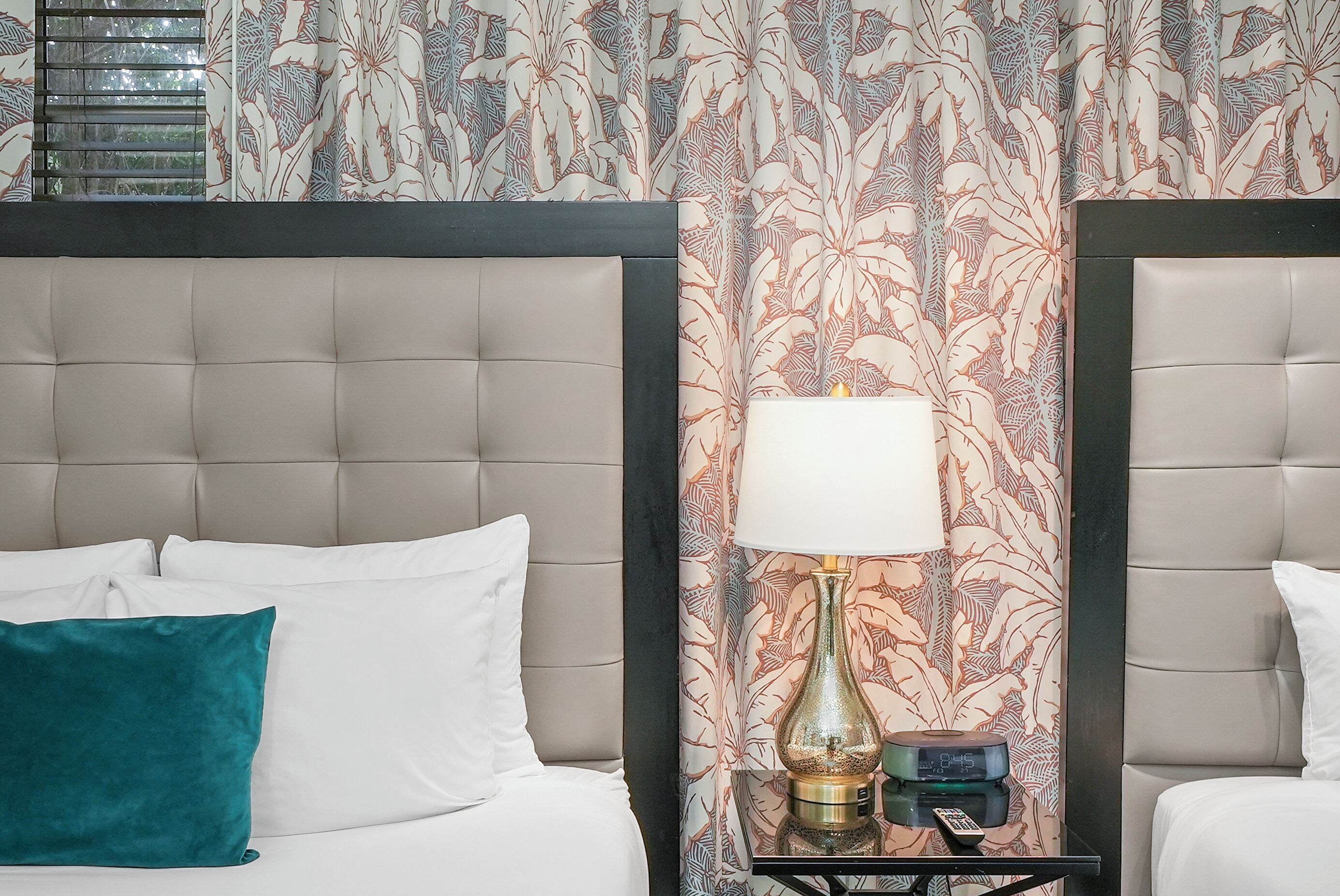 A hotel room with a tufted headboard, a bed with white and teal pillows, a bedside table with a lamp and a clock, and a floral-patterned curtain.