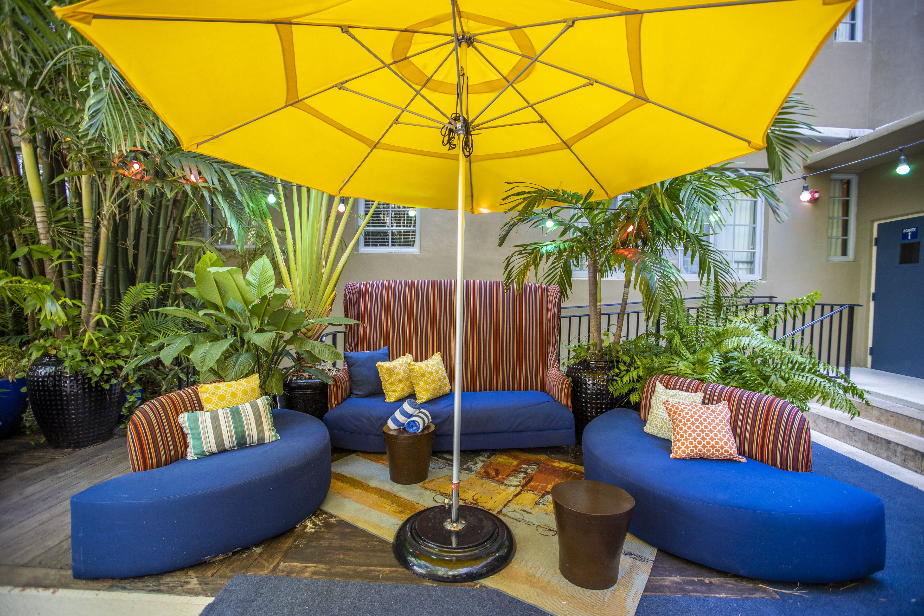 Un espace extérieur aménagé avec des sièges bleus, un parasol jaune, des plantes en pot et un rideau à rayures.