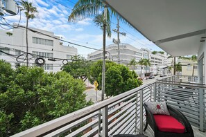 A balcony with a wicker chair and red cushion, overlooking a street with buildings and palm trees. - Riviera Hotel South Beach (Miami Beach)